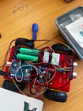 Top-down view of a recycled-parts robot car under construction, with battery, breadboard and jumper wires on a classroom desk.